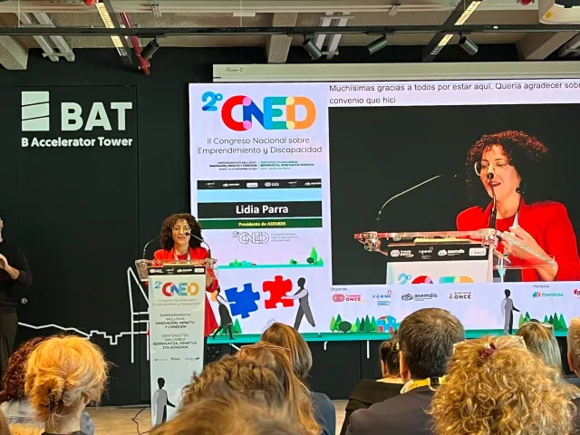 Lidia Parra, presidenta de ASEMDIS, interviene desde el atril del II CNED en la BAT de Bilbao, con su imagen ampliada en la pantalla