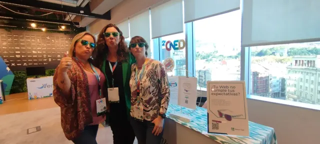 Tres asistentes posan con gafas de sol junto a un stand en el espacio de exposición del II CNED