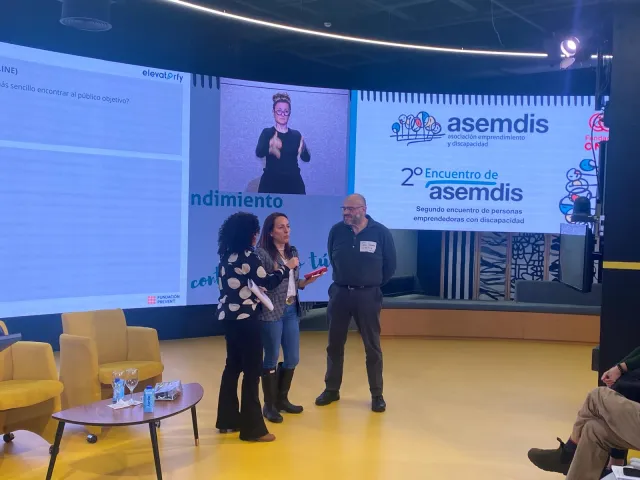 Tres personas en el escenario durante la sesión de Elevatorfy, con una intérprete de lengua de signos en la pantalla y el cartel del 2.º Encuentro de ASEMDIS