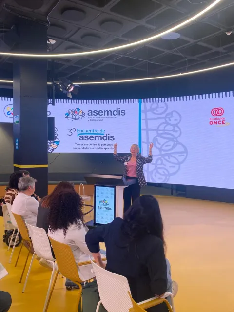 Una ponente da la bienvenida desde el escenario del 3.º Encuentro de ASEMDIS ante el público sentado