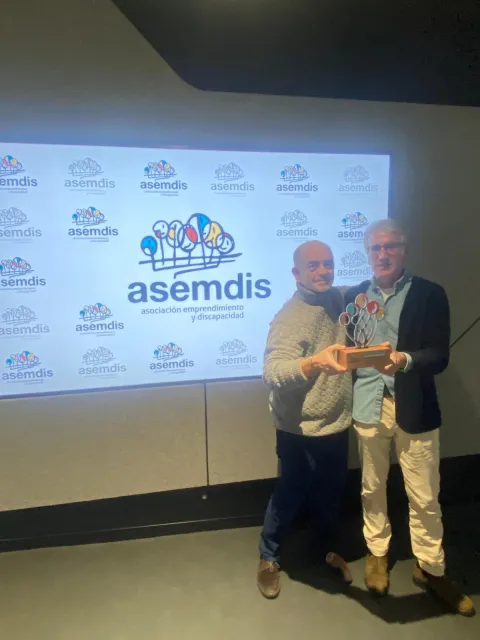 Dos personas sostienen un reconocimiento de ASEMDIS ante un photocall con el logotipo de la asociación